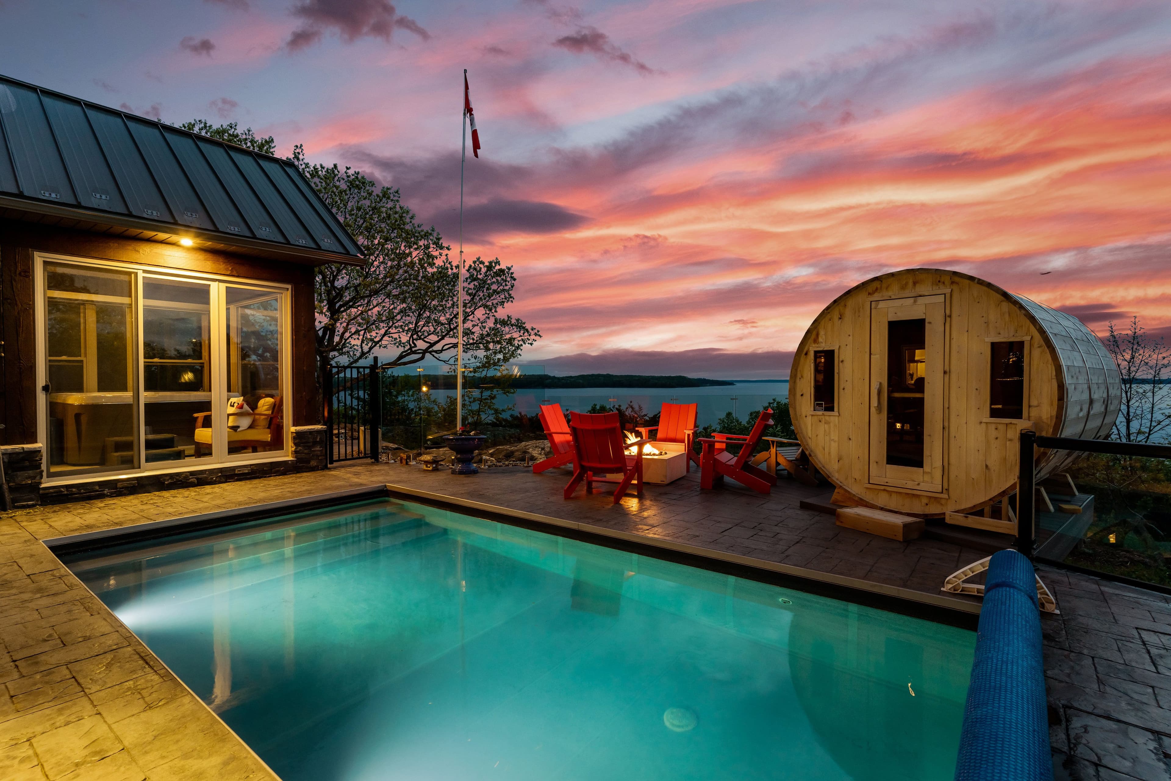 A poolside patio with red Muskoka chairs, a fire pit, and a barrel sauna overlooking the lake at sunset.