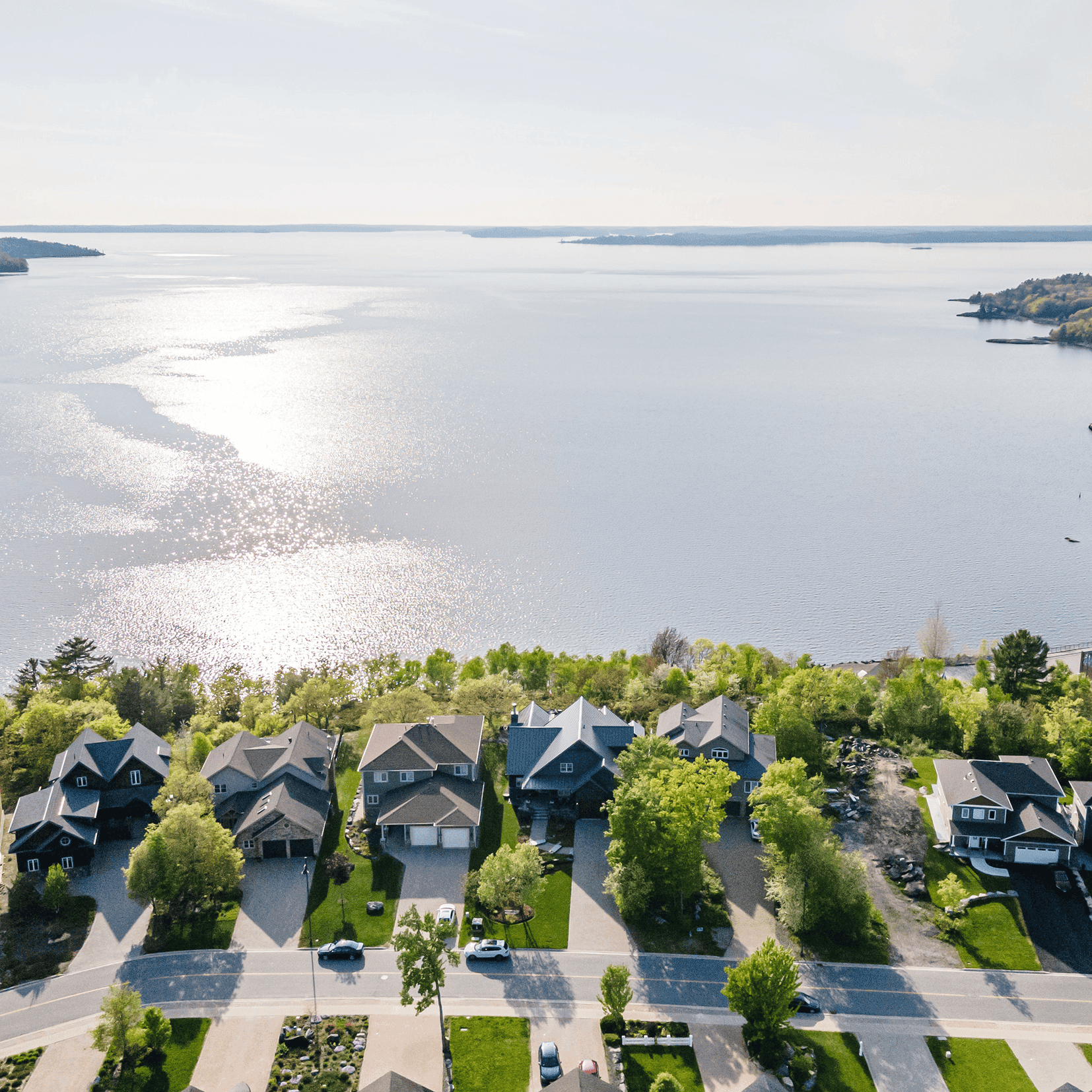 Aerial view of a row of suburban houses with green lawns facing a large, sparkling body of water.